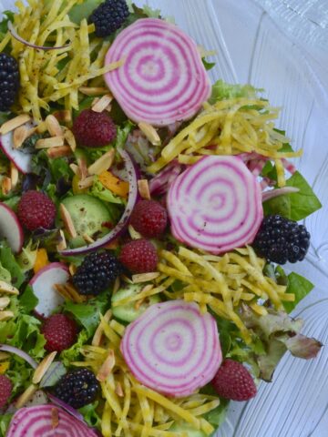 A glass bowl with Garden Harvest Jewelled Salad containing sliced candy cane beets, grated golden beets, raspberries, blackberries and a variety of lettuce.