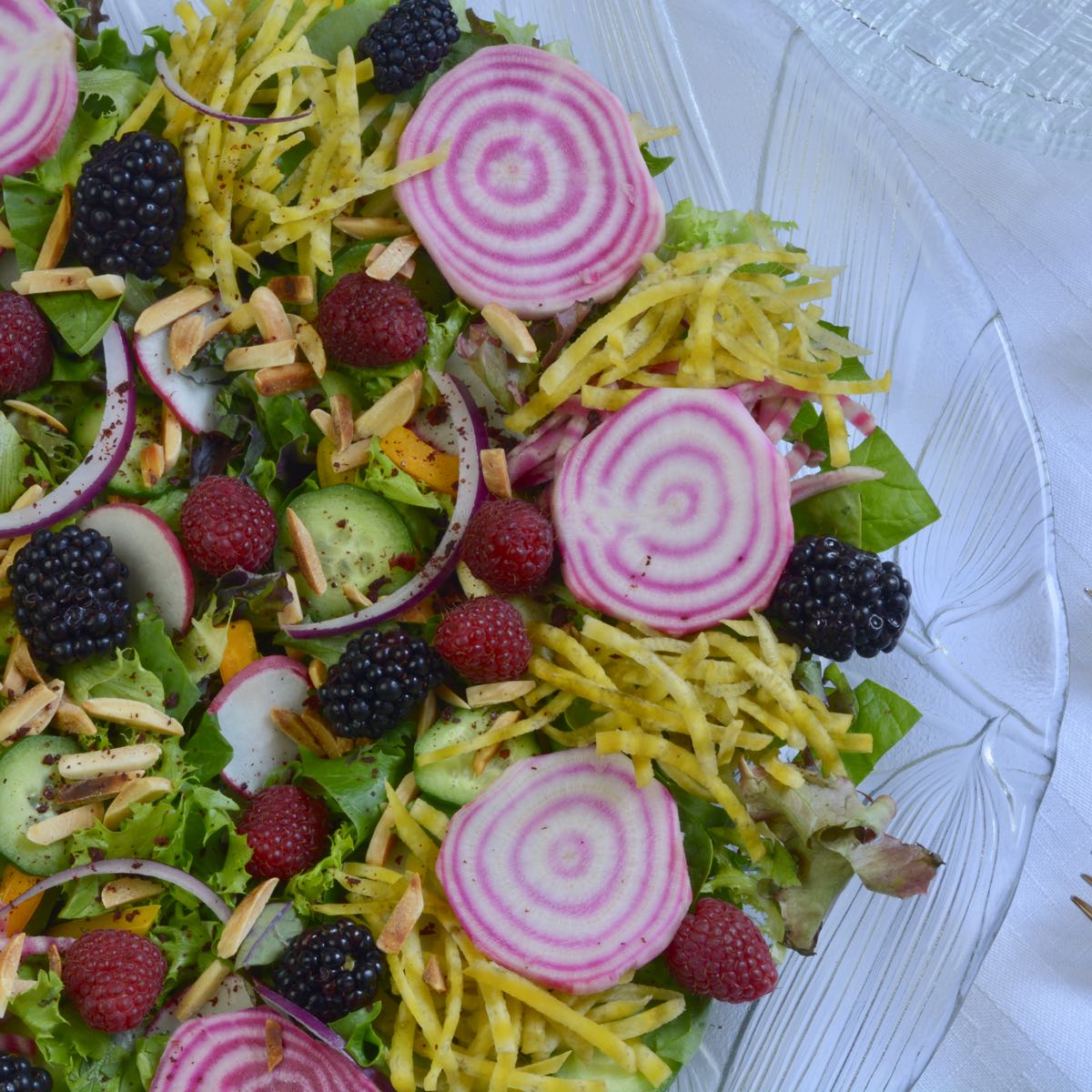 A glass bowl with Garden Harvest Jewelled Salad containing sliced candy cane beets, grated golden beets, raspberries, blackberries and a variety of lettuce.
