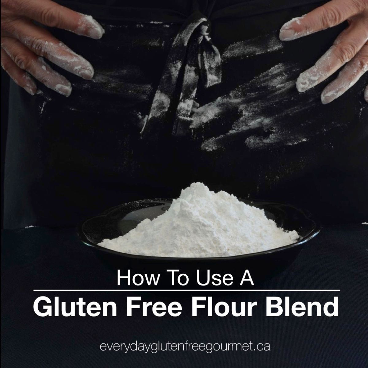 Two floured hands, wiping flour on a black apron from a bowl of flour in front of them.