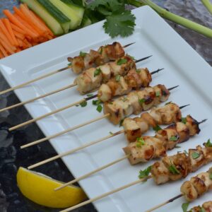 A platter with skewers of Lemongrass Chicken surrounded by lemongrass, carrots, cucumber and lemon wedges.