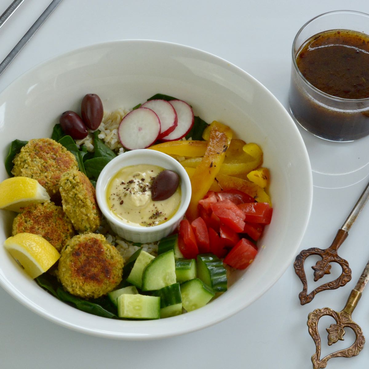 A white bowl with rice, falafel balls, hummus and chopped vegetables with pomegranate dressing on the side.