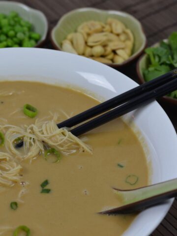 A bowl of Noodle Soup with Peanut Sauce surrounded by optional toppings; peanuts, green onions ad cilantro.
