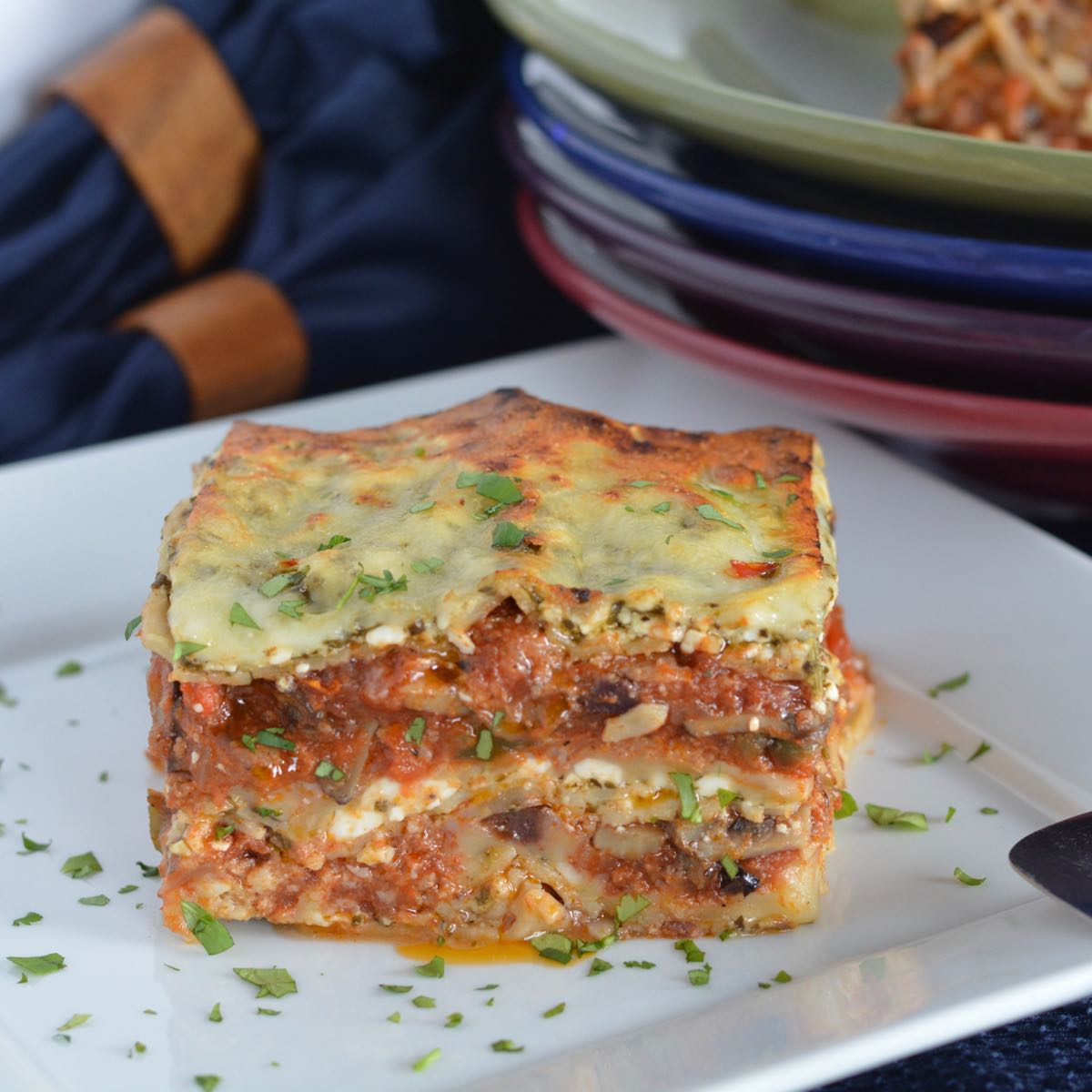 A piece of pesto lasagna sprinkled with parsley sitting beside a stack of colourful bowls.