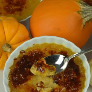 A dish of Pumpkin Creme Brulee with the first bite on the spoon surrounded by orange and white mini pumpkins.