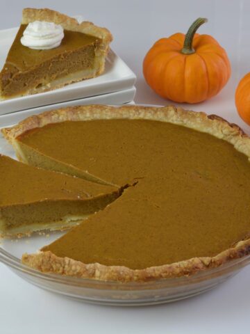 A whole gluten free Pumpkin Pie with one piece placed on a plate and 2 mini pumpkins beside it.