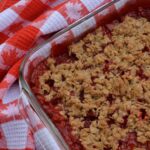 A pan of Rhubarb Strawberry Crisp surrounded by a tea towel with a maple leaf pattern.