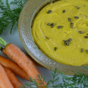 Roasted Carrot Hummus in a round dish sprinkled with toasted pumpkin seeds and surrounded by garden carrots.