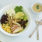 A bowl filled with Santa Fe Salad; blackened chicken slices, avocado, corn, beans, feta cheese and dates sprinkled with toasted pumpkin seeds.