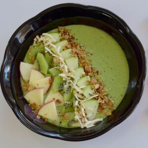 A green smoothie in a black bowl garnished with cut up apple and kiwi, sprinkled with granola; and a few wedges of red and green apple beside the bowl.