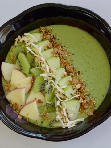 A green smoothie in a black bowl garnished with cut up apple and kiwi, sprinkled with granola; and a few wedges of red and green apple beside the bowl.