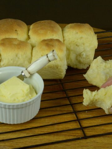A wire rack with 7 dinner rolls still together and one dinner roll broken open with a dish of butter., tasting soft dinner rolls