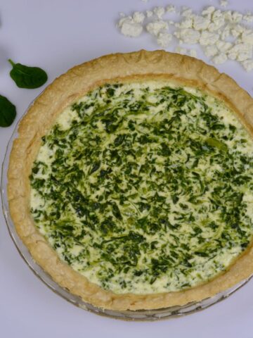 A cooked Spinach Feta Quiche surrounded by fresh spinach, crumbled feta and two eggs.