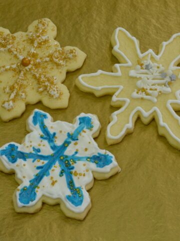 Three gluten-free snowflake sugar cookies decorated with royal icing and sprinkles on gold tissue paper.