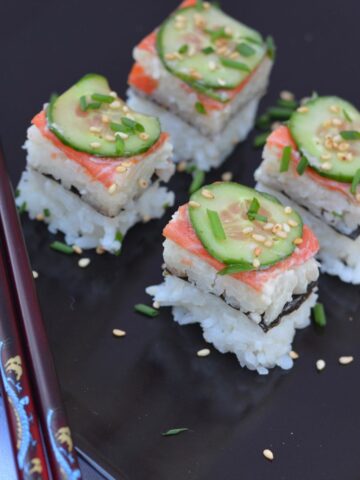 A black tray of smoked salmon sushi pizza cut in squares and sprinkled with snipped chives and sesame seeds.