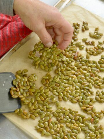 A baking sheet of Candied Sweet and Salty Pumpkin Seeds with a flipper and a hand breaking a piece apart.