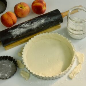 An uncooked Gluten free tart pastry on a counter with the rolling pin and fresh peaches.