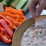 A cracker being dipped into a dish of warm Cranberry Dip surrounded by colourful raw veggies.