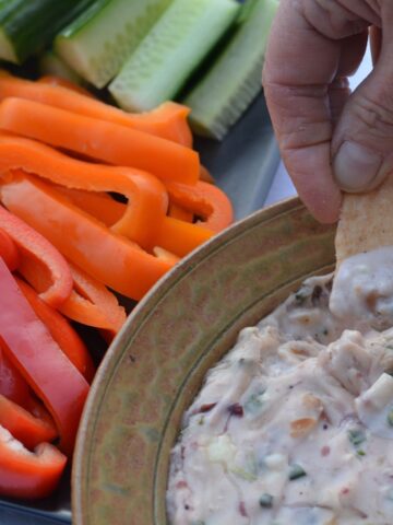 A cracker being dipped into a dish of warm Cranberry Dip surrounded by colourful raw veggies.