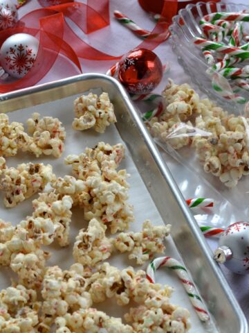 A pan with clumps of White Chocolate Candy Cane Popcorn beside gift bags, candy canes and ribbon.