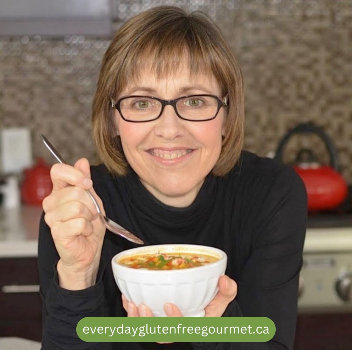 Cinde in her kitchen holding a bowl of soup with a spoon in hand.