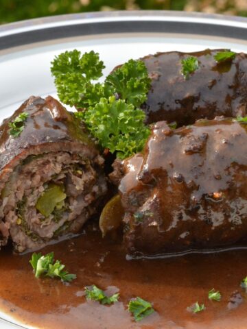 A plate with 3 rolls of Beef Rouladen covered in a rich red wine gravy and sprinkled with parsley.