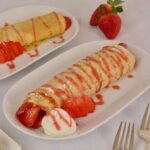 Two plates each with a gluten free crepe filled with whipped cream, drizzled with Strawberry Sauce and garnished with fresh strawberries.