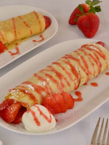 Two plates each with a gluten free crepe filled with whipped cream, drizzled with Strawberry Sauce and garnished with fresh strawberries.