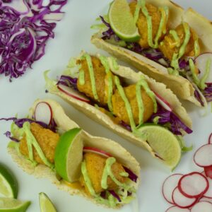 A platter of fish tacos drizzled with avocado crema and topped with lime wedges.