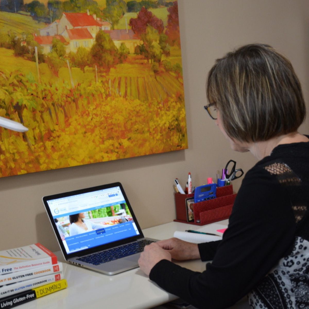 Cinde at a desk looking at a laptop with a stack of books beside her for learning about the gluten free diet.