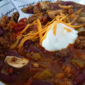 Close up of a bowl of Homemade Chili topped with a blog of sour cream and sprinkled with shredded cheddar cheese.