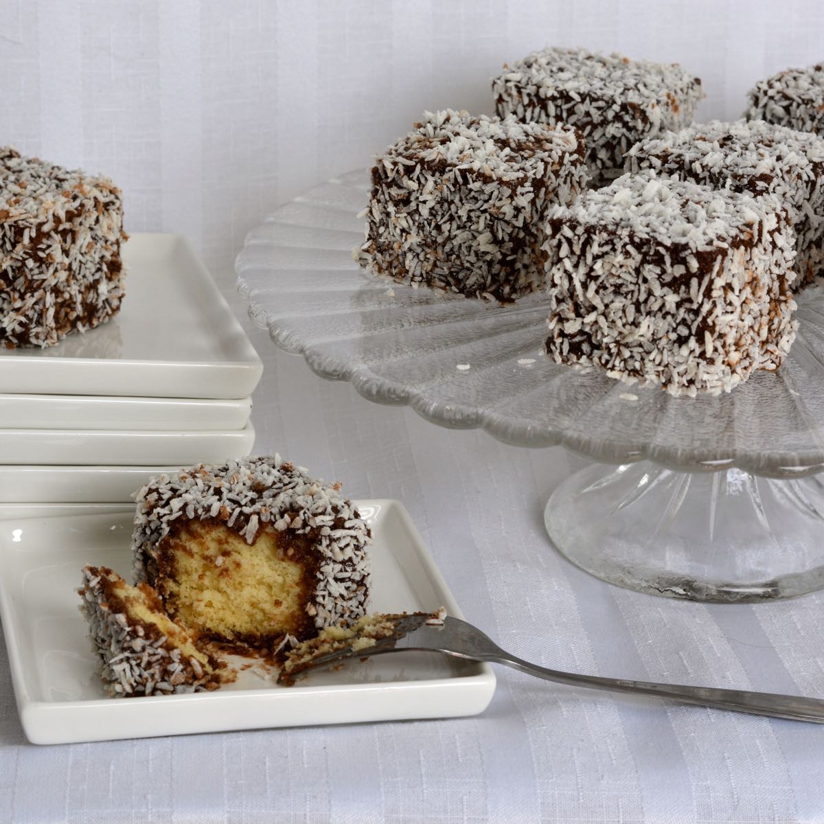 A pedestal tray with squares of gluten free lamingtons, with small plates beside it and one lamington cut into with a fork.