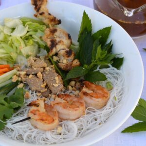 A white bowl filled with rice noodles, grilled shrimp, chicken and pork, shredded lettuce and fresh herbs with a dish of nuoc cham dipping sauce beside it.