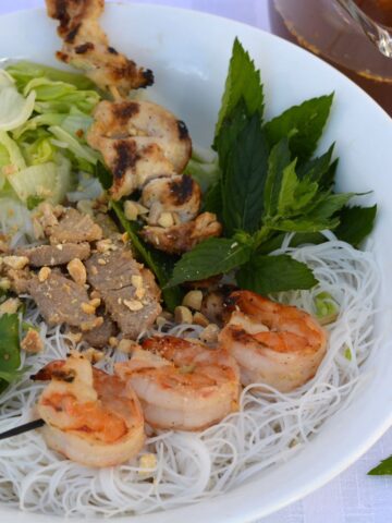 A bowl filled with rice noodles, grilled shrimp, chicken and pork, shredded lettuce and fresh herbs with a dish of nuoc cham dipping sauce beside it.