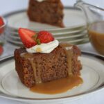 Aa piece of Gluten Free Sticky Date Pudding on a plate with caramel sauce dripping down the sides topped with whipped cream and a strawberry.