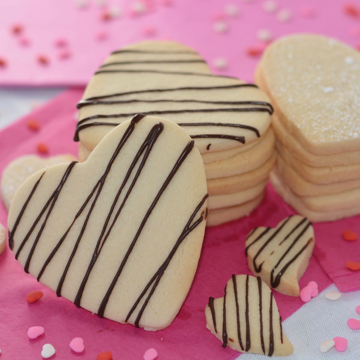 A stack of heart shaped gluten free sugar cookies drizzled with chocolate sitting on pretty pink paper surrounded by tiny heart shaped sprinkles.