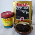 A tub of Tamarind concentrate beside a package of tamarind paste, with a small dish in front showing the paste.