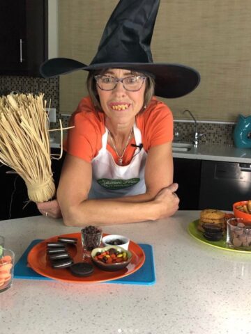 Cinde in her kitchen wearing an orange shirt, witch hat and crooked teeth holding a broom. Bowls of Halloween candy in front of her.