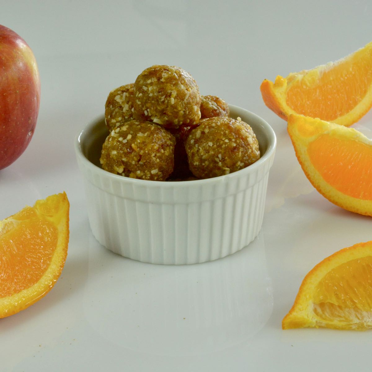 A small dish of Coconut Date Energy Balls surrounded by orange wedges and an apple.