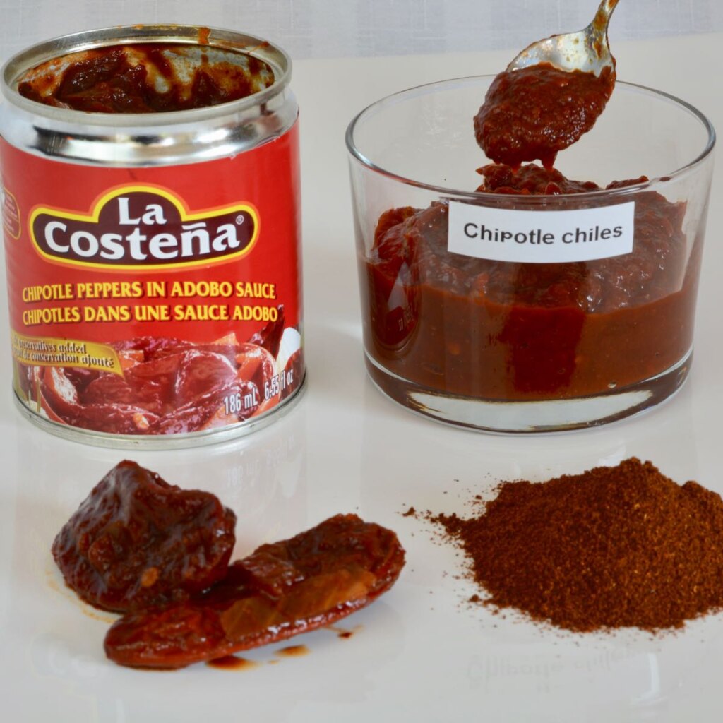 A small tin of Chipotle Chiles in Adobo Sauce with a dish of them beside it and a mound of chipotle chile powder beside it.