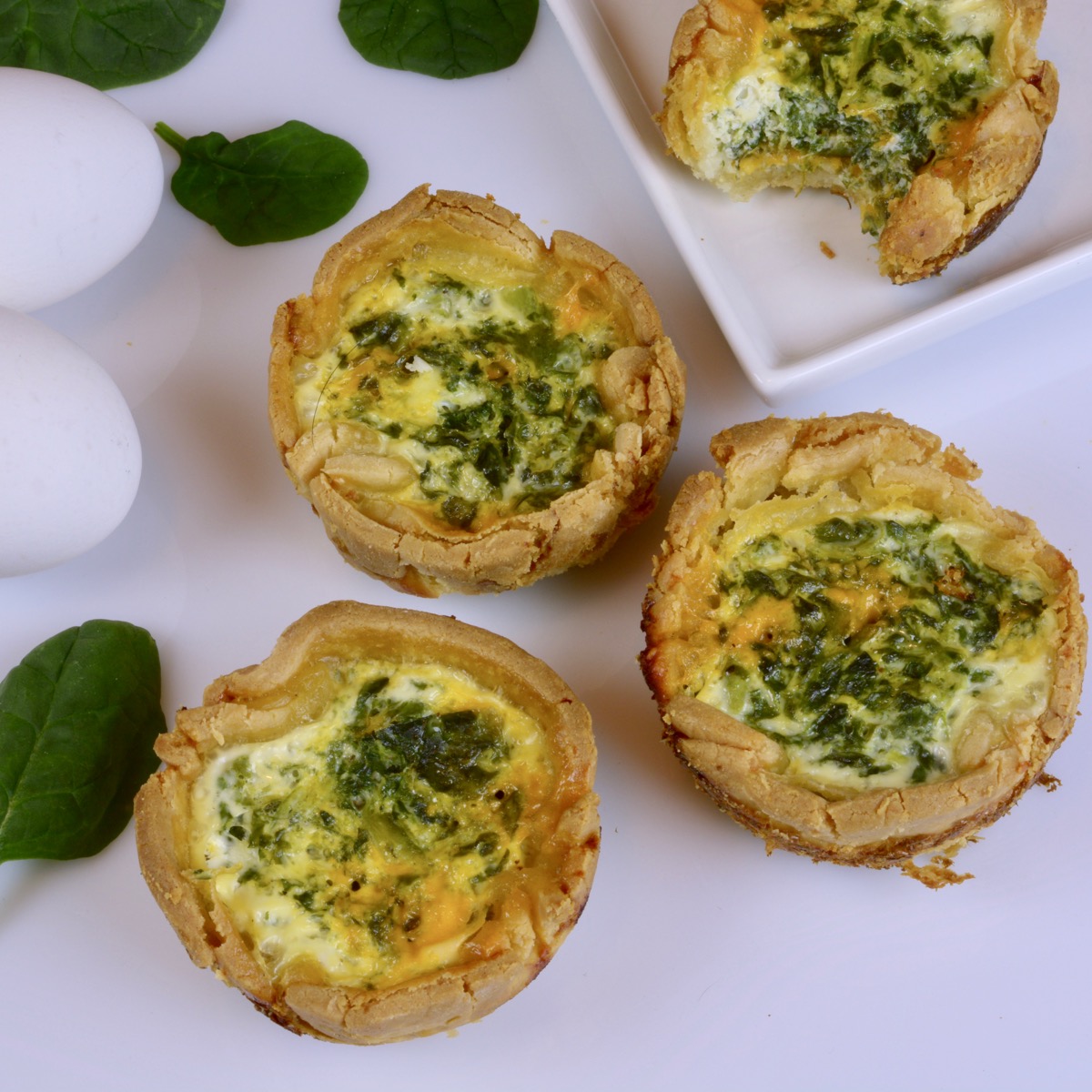 Three spinach and feta quiche tarts surrounded by white eggs and spinach leaves.