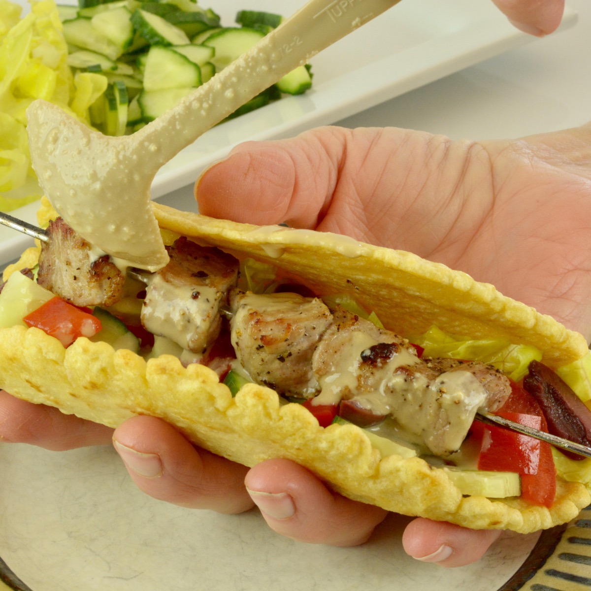 Someone holding a gluten free chickpea flour wrap filled with pork souvlaki, tomato and cucumber with a tahini sauce being poured on top.