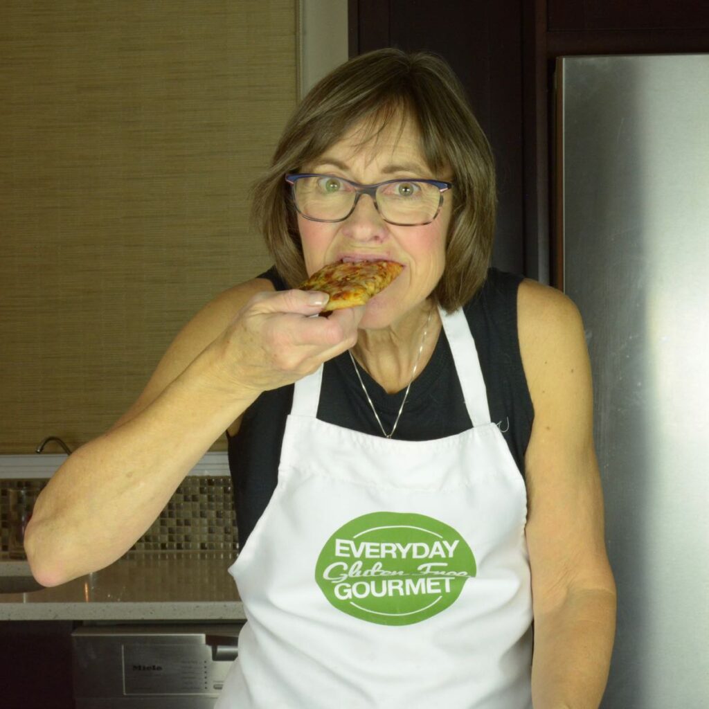 Cinde wearing her Everyday Gluten Free Gourmet apron, standing in her kitchen and biting into a piece of pizza.