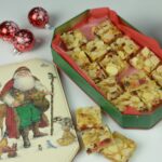 A holiday tin filled with gluten free Fruitcake Squares surrounded by Christmas festive balls and more squares.