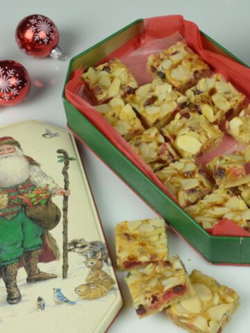 A holiday tin filled with gluten free Fruitcake Squares surrounded by Christmas festive balls and more squares.