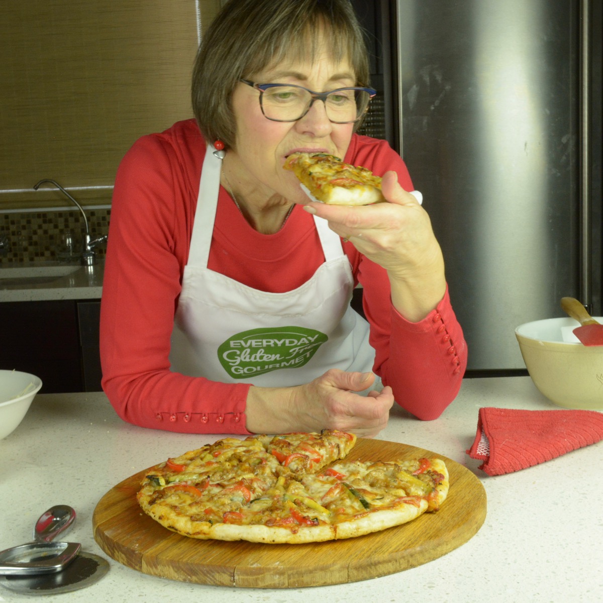 Cinde leaning on the counter in front of a cooked pizza, taking a bite out of the piece in her hand.