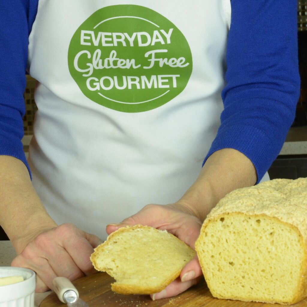 Someone holding a warm slice of gluten free millet bread, beside the whole loaf it was cut from, covered with butter.