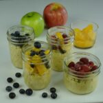 Four jars of overnight oats topped with oranges, blueberries and raspberries surrounded by whole apples and a few blueberries.