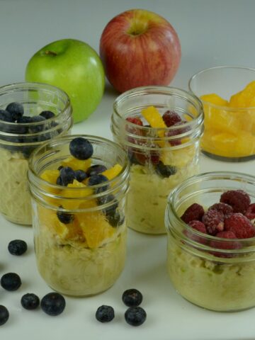 Four jars of overnight oats topped with oranges, blueberries and raspberries surrounded by whole apples and a few blueberries.