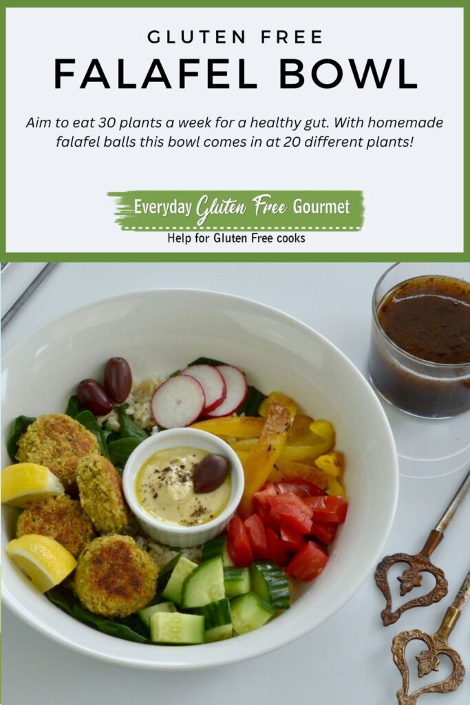 A bowl with rice, falafel balls, hummus, chopped cucumber, roasted peppers, radish, olives and spinach. There is a bowl of pomegranate dressing on the side.