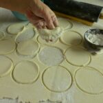 A hand holding one perogy above a counter covered in circles of dough ready to make more perogies.